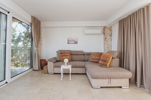 Una sala de estar moderna con un sofá beige, una mesita auxiliar blanca y vistas a un espacio exterior a través de un ventanal.