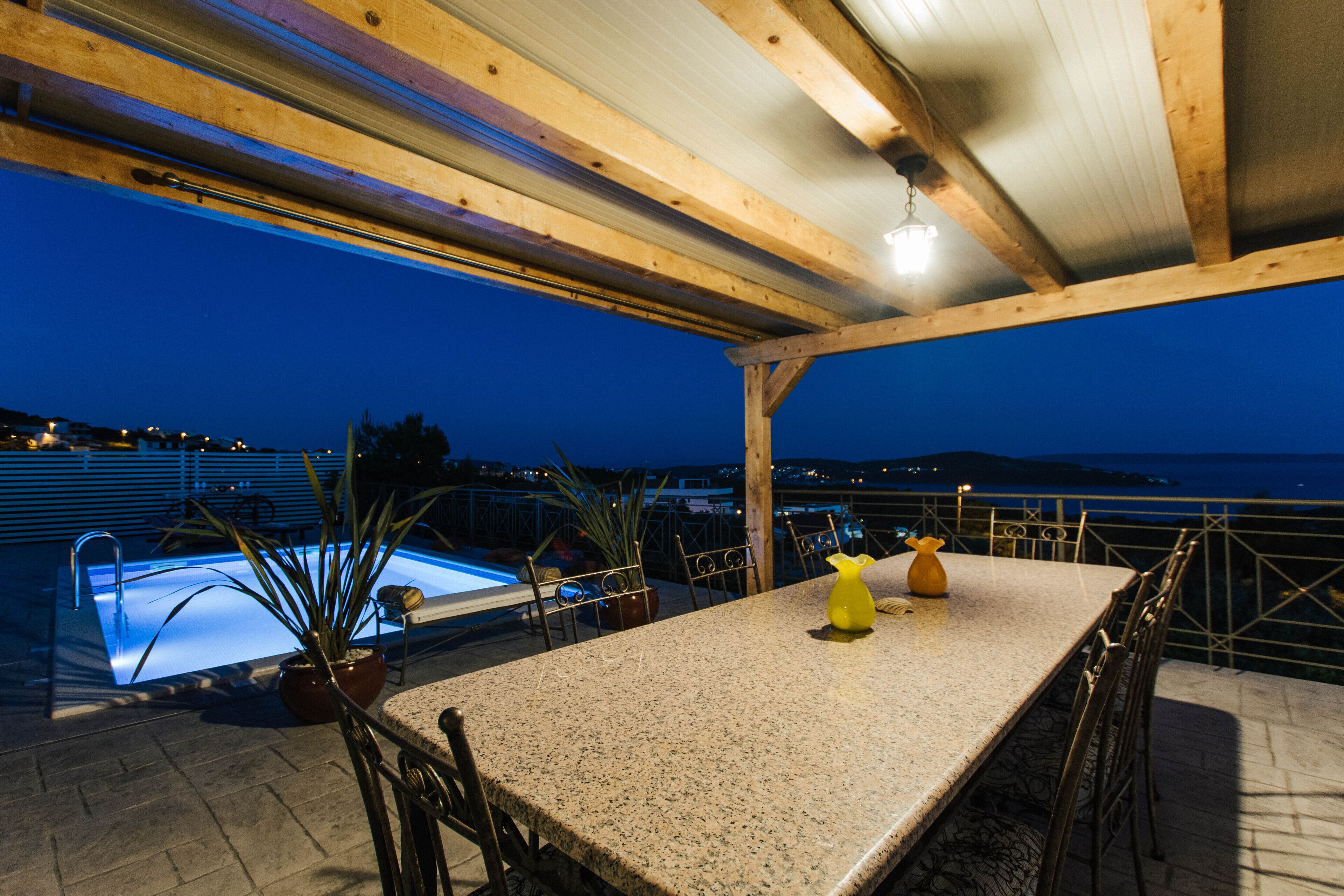 An outdoor pool area with a table and chairs, surrounded by a wooden pergola structure.