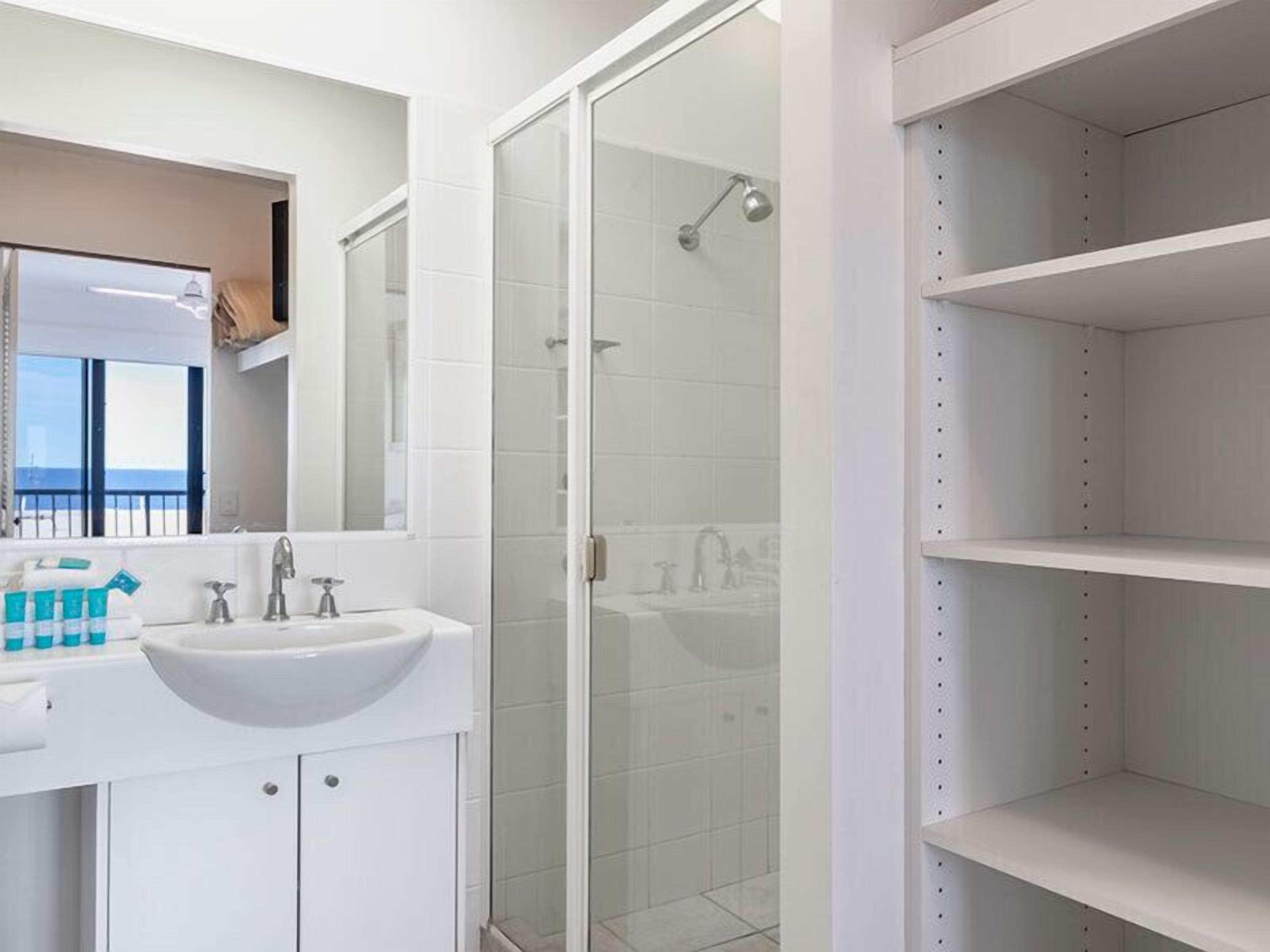 Un baño moderno con ducha envuelta en vidrio, un espejo grande y un lavamanos blanco con artículos de aseo.