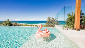 Ein klares, blaues Schwimmbecken mit einem rosa Flamingo-Schwimmpfad, mit Blick auf einen Strand mit Meereswellen und einem wolkenlosen Himmel.