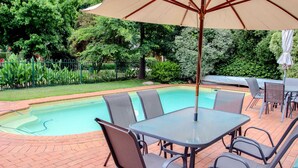 En swimmingpool med fliseterrasse, omgivet af en have med forskellige planter og et hegn.