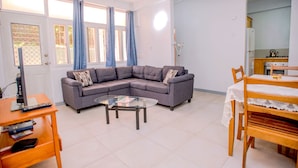 A living room with a sectional sofa, a glass coffee table, a TV on a wooden stand, and a dining area with a table and chairs.