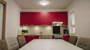 Cocina pequeña con armarios rojos, estufa y lavamanos. En primer plano, una mesa de comedor con sillas.
