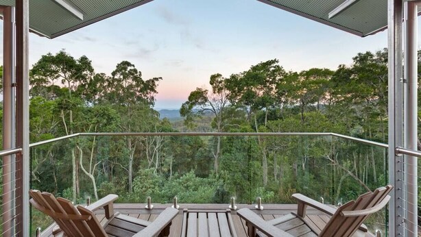 A balcony with wooden chairs and a glass railing overlooking a forested area.
