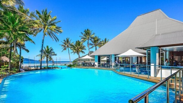 A large, clear blue pool with a deck area, surrounded by palm trees and a building with a grey roof.