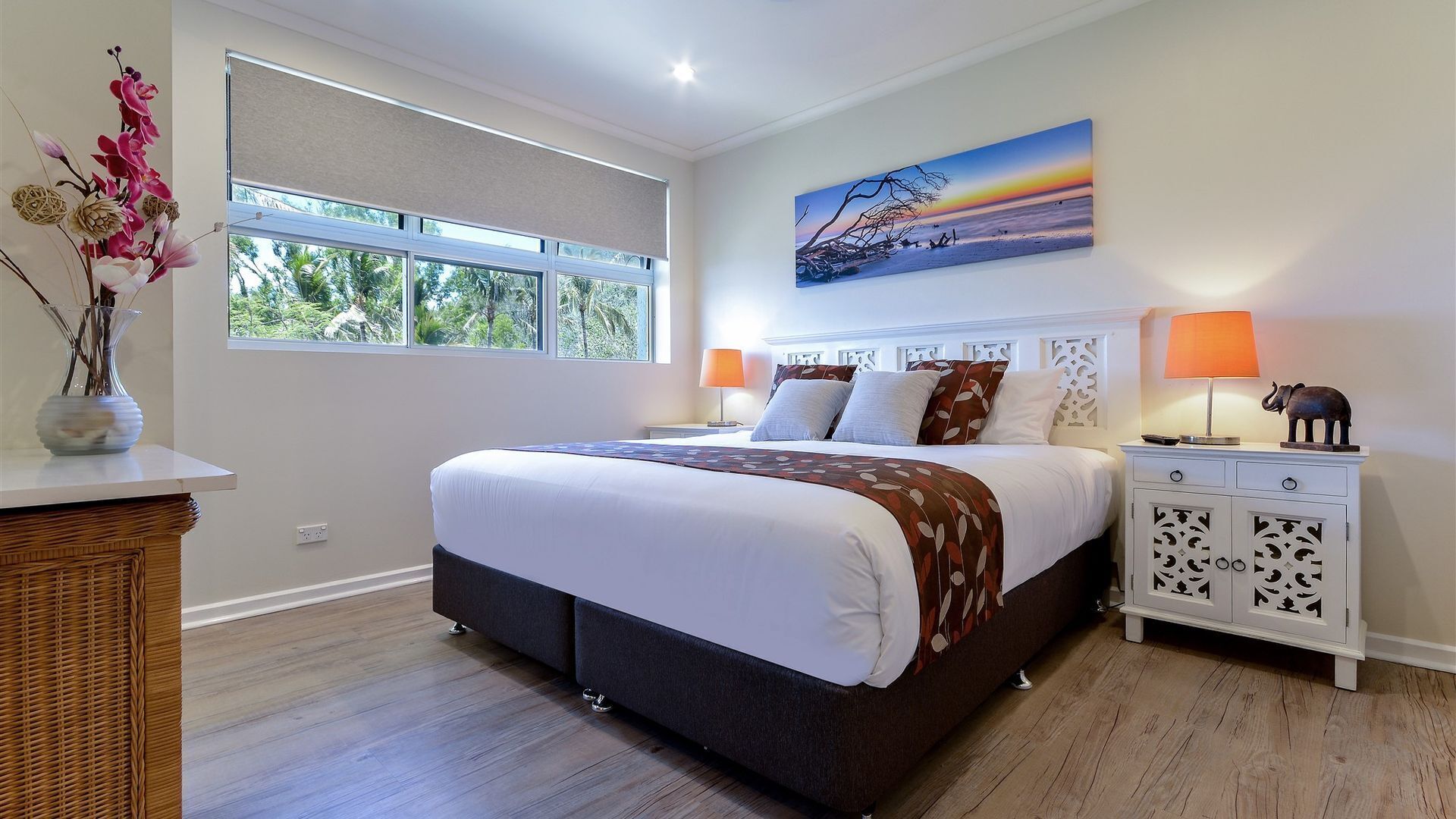 Une chambre avec un grand lit, deux tables de chevet, une commode, une fenêtre donnant sur un paysage verdoyant et un tableau accroché au mur.