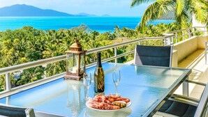 A table with a bottle of champagne, two glasses, and a plate of shrimp and olives, overlooking a tropical landscape with a view of the ocean and mountains.
