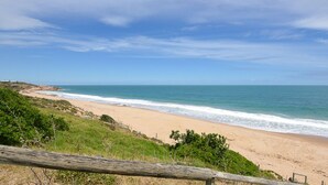 Une plage avec une clôture en bois au premier plan, une rive sablonneuse et l’océan sous un ciel dégagé.