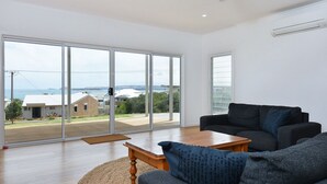 Un salon spacieux doté de grandes portes-fenêtres coulissantes, d’un canapé gris foncé, d’une table basse en bois et d’une vue sur un quartier résidentiel avec des maisons et de la verdure.