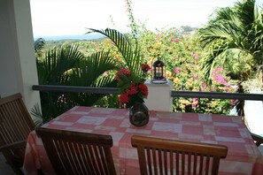 Ein Balkon mit Tisch und St ühlen, der auf einen üppigen Garten und den Blick aufs Meer blickt.