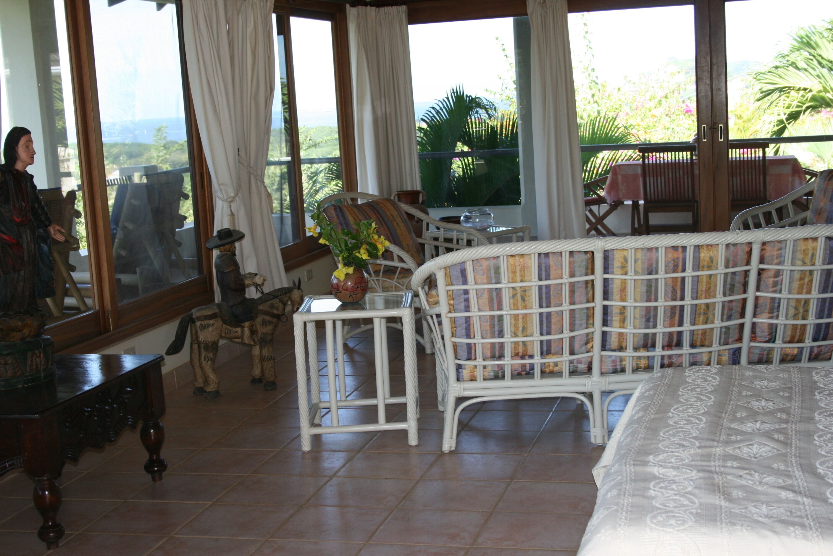 A room with a bed, a white wicker sofa, a small table with a vase, and a chair. There are curtains on the windows, and a view of greenery outside.