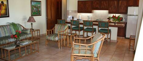 Una sala de estar con sofá, mesa de centro y sillas, una cocina con armarios de madera y electrodomésticos, y una puerta que da a otra habitación.
