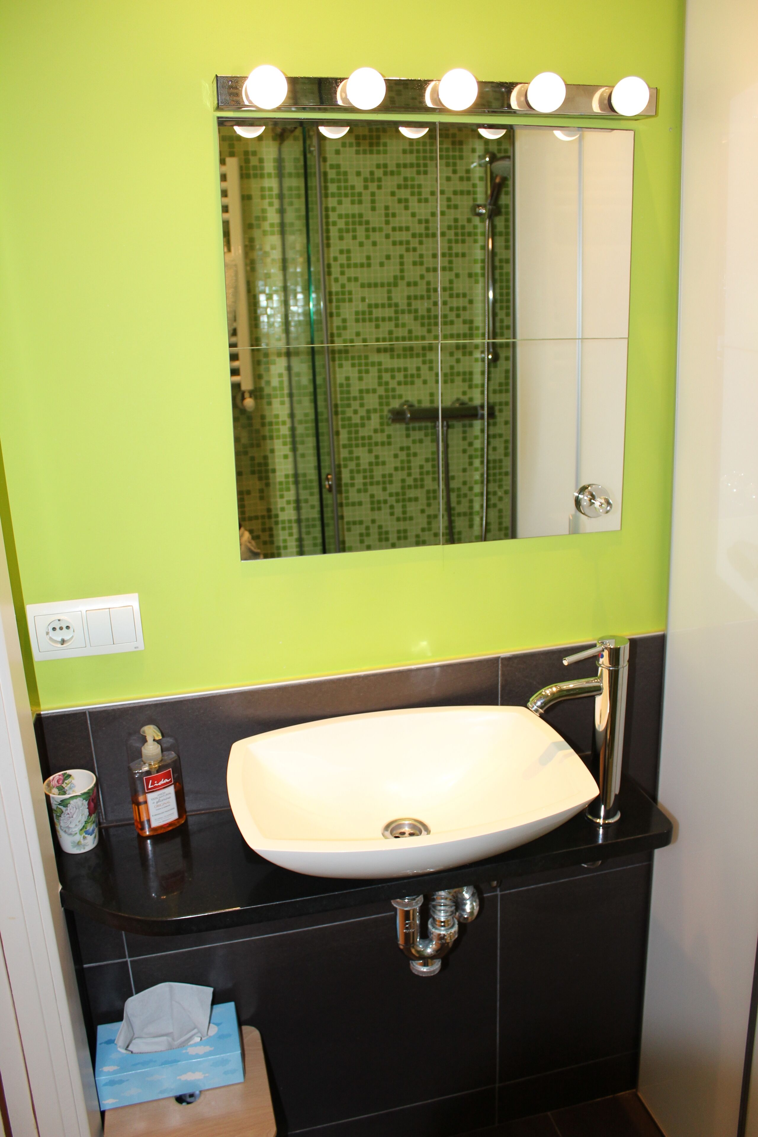 Een badkamer met een witte wastafel, een spiegel met verlichting en een douche met groene tegels.