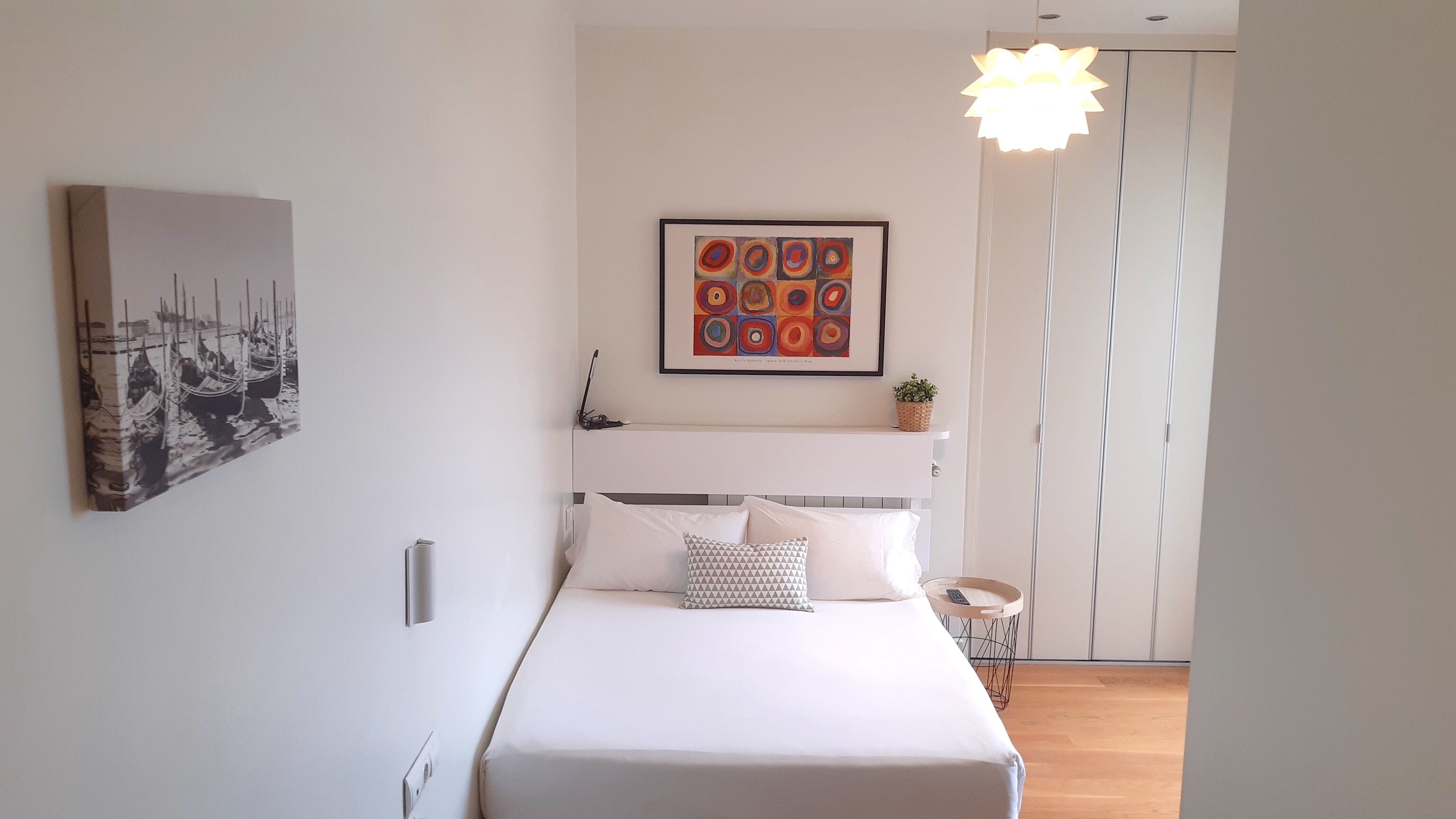 A modern bedroom with a bed, a framed abstract painting, a small plant, and a unique light fixture.