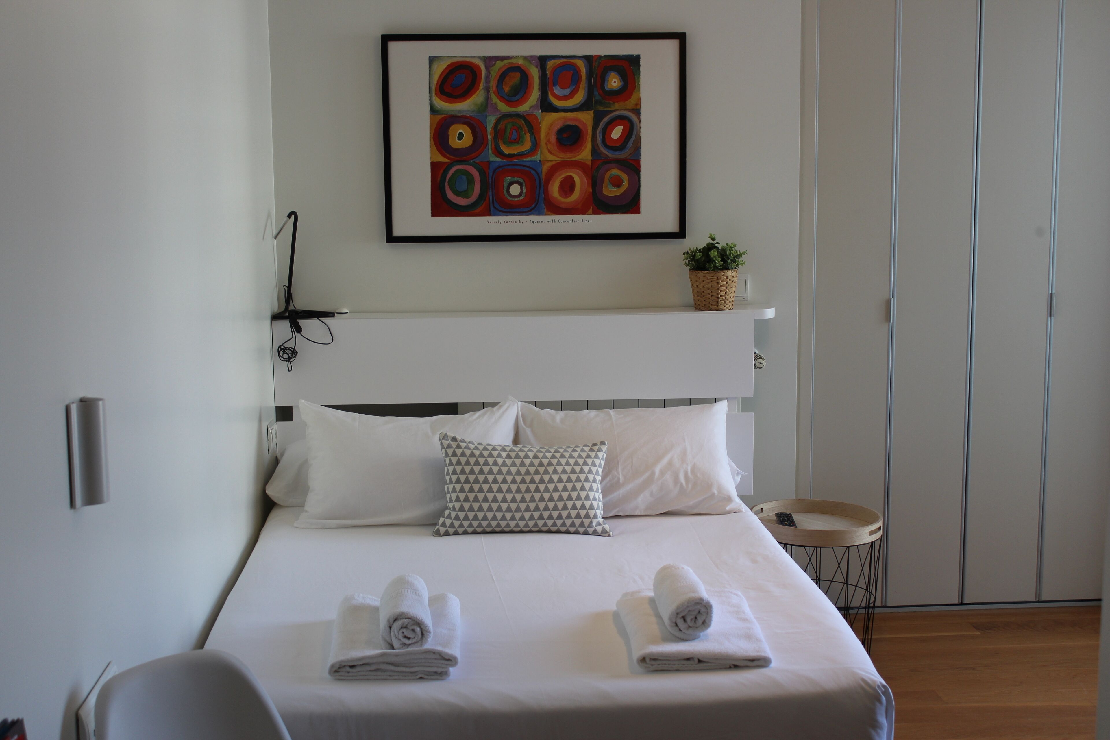 A neatly made bed with white linens and two rolled towels, a geometric patterned pillow, a framed abstract painting above the bed, a small plant on a shelf, and a bedside table with a tray.