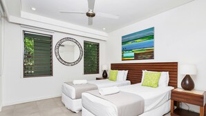 Een moderne hotelkamer met twee bedden, een plafondventilator, een groot raam met jaloezieën, een ronde spiegel en een schilderij aan de muur.