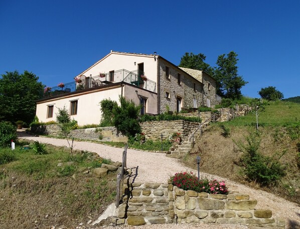 Une maison en pierre à deux étages, dotée d’un balcon, entourée d’un chemin de gravier et de murs en pierre.