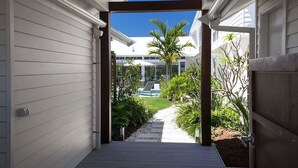 A covered outdoor area with a pathway leading to a pool, surrounded by greenery and a clear blue sky. - White Sands Beach House (Marcoola)