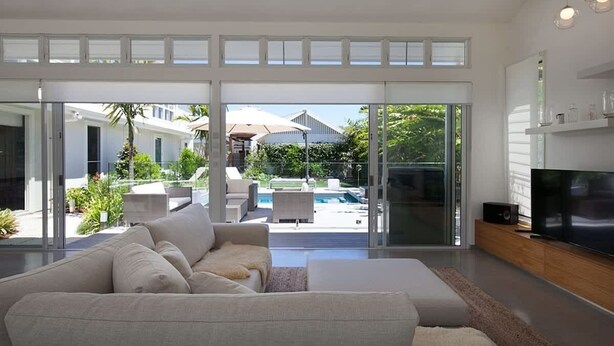 A modern living room with a large sofa, a flat-screen TV, and a pool view. - White Sands Beach House (Marcoola)