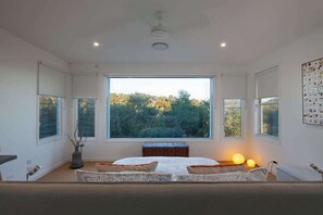 Un dormitorio con un ventanal grande, una cama con ropa de cama estampada, una mesita de noche con lámparas y un ventilador de techo.