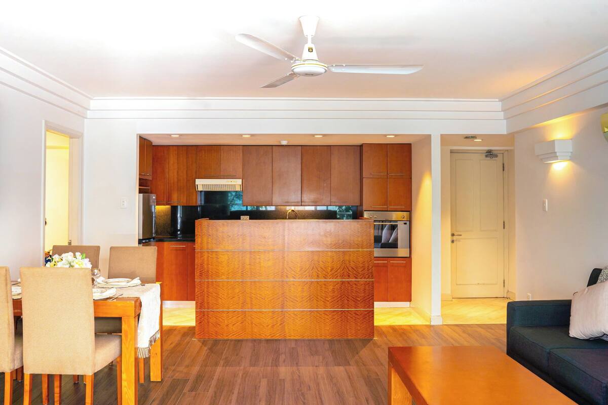 A modern living room with a dining area, wooden cabinets, a ceiling fan, and a kitchen in the background.