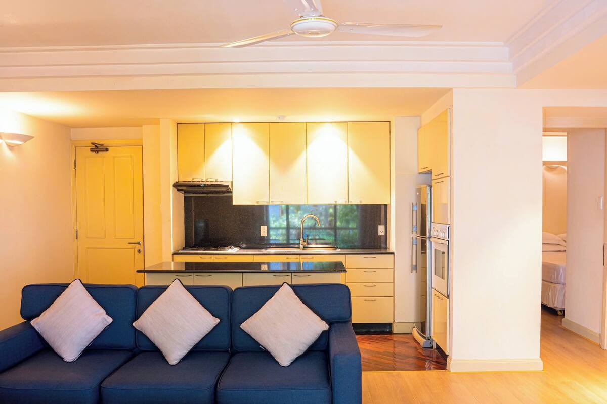 A modern kitchen with a built-in oven and refrigerator, a blue sofa with cushions, and a ceiling fan.