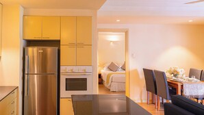 A modern kitchen with stainless steel appliances, a dining area with a table set for two, and a bedroom visible through an open doorway.