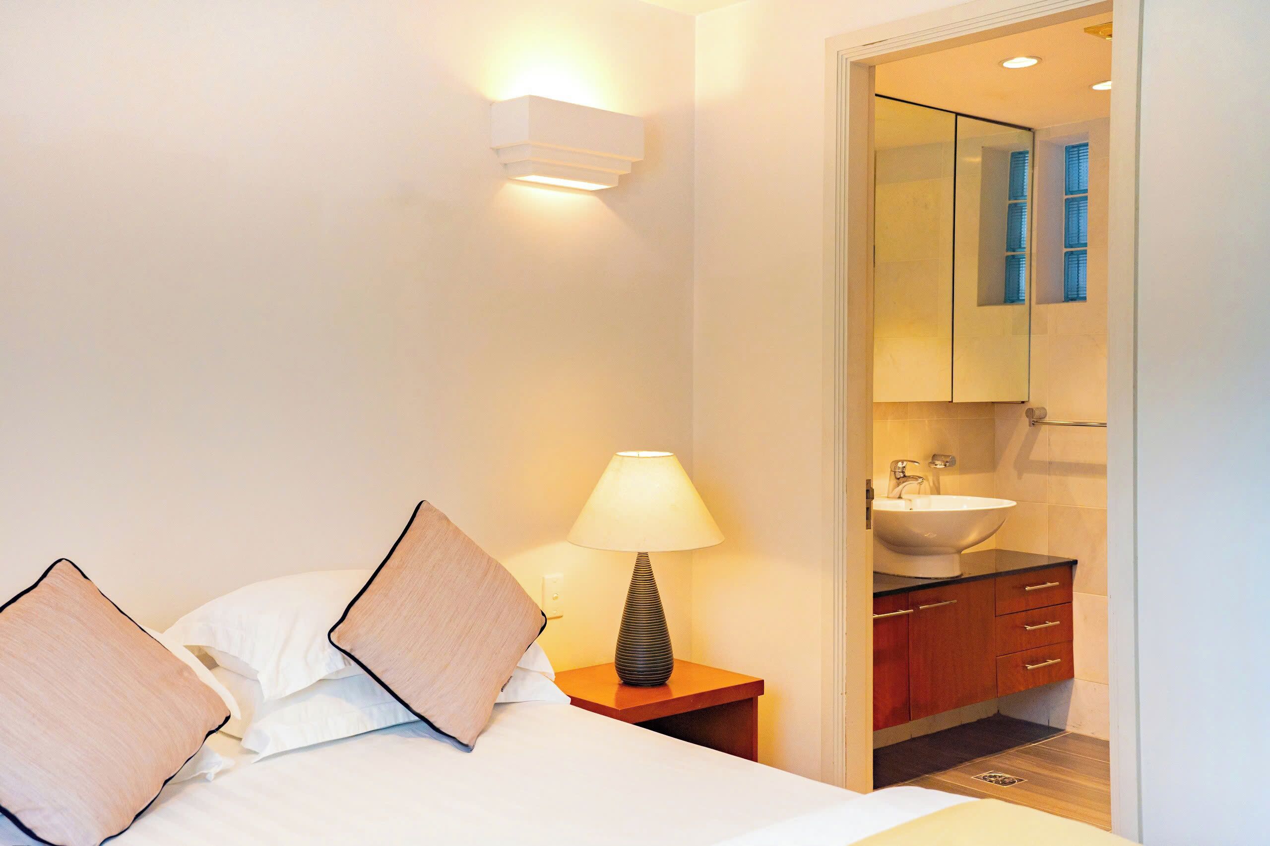 A hotel room with a bed, a bedside lamp, a nightstand, and a bathroom visible through an open door.