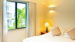 A hotel room with a bed, a nightstand with a lamp, a window with curtains, and a view of buildings and trees outside.