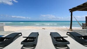 Un espace de détente aménagé au bord de la plage, avec trois chaises longues orientées vers l’océan, une pergola en bois et une allée piétonne en béton.
