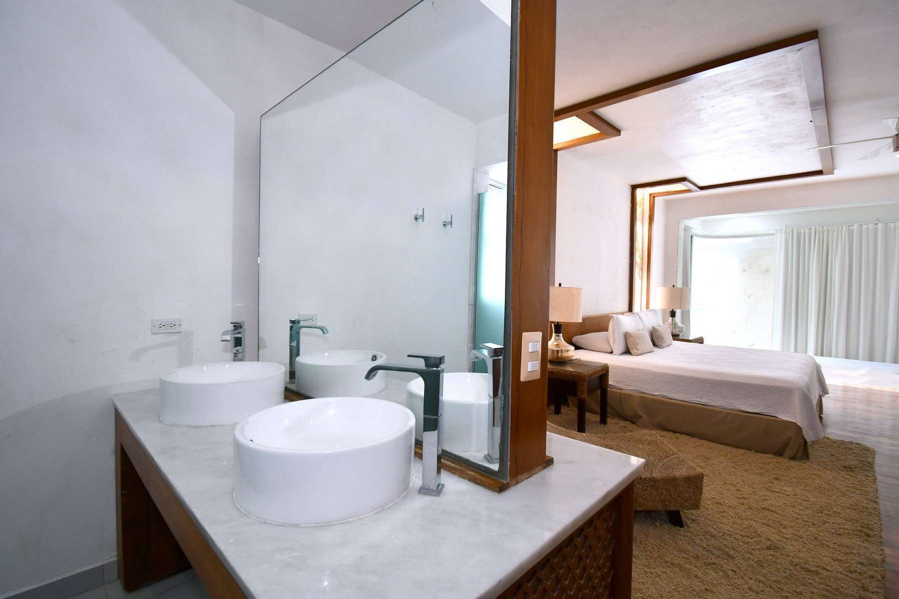 Een hotelbadkamer met twee wastafels, een grote spiegel en een bed in de aangrenzende kamer.