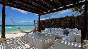 Une terrasse au bord de la plage, avec un hamac, un canapé agrémenté de coussins et une table en verre.