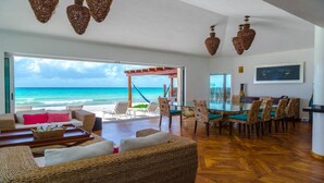 Un vaste espace de vie avec une grande porte vitrée donnant sur la plage, des meubles en osier et un coin repas avec une table et des chaises en verre.