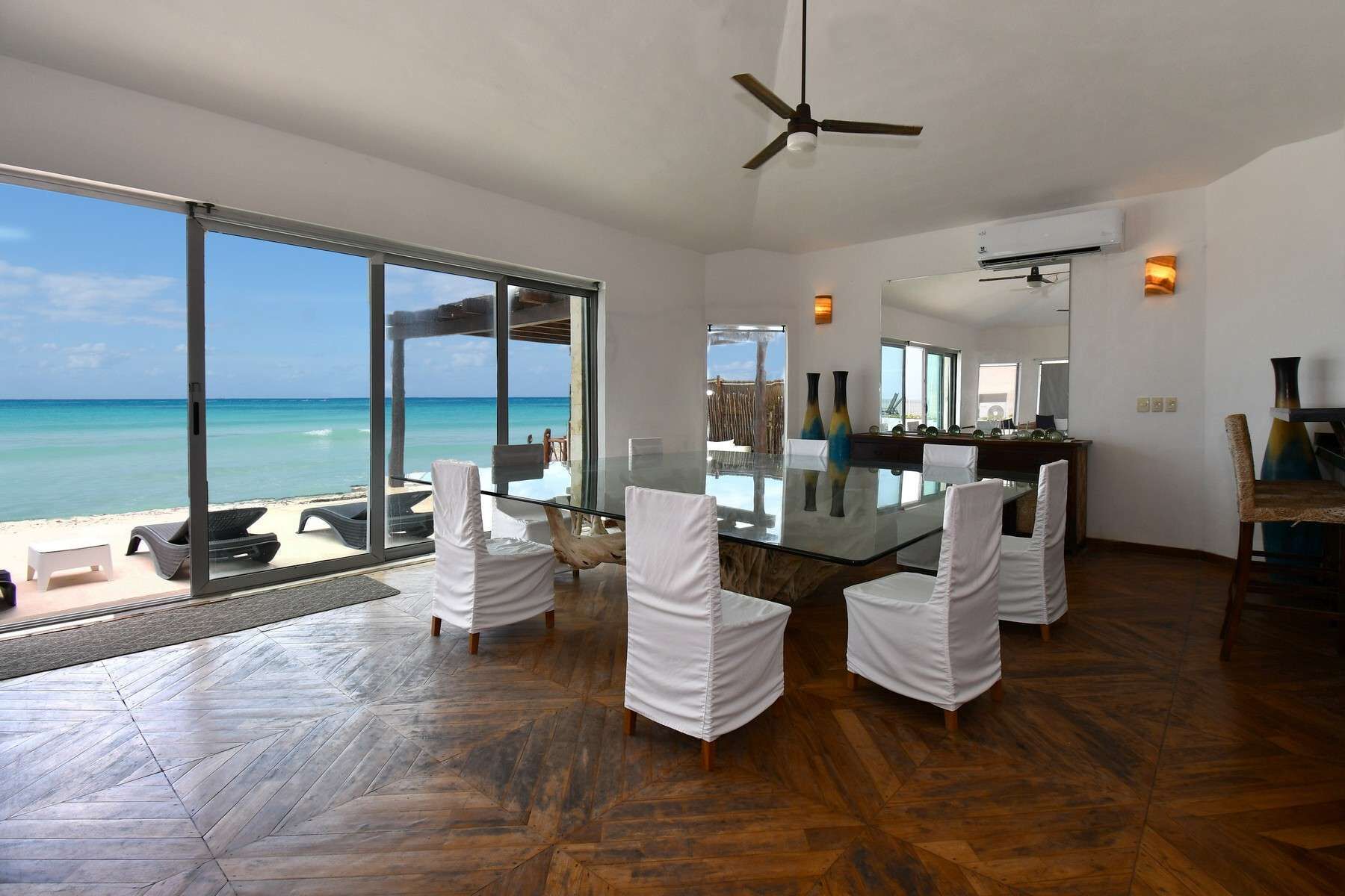 Un vaste espace dédié à la salle à manger, avec une table en verre et des chaises blanches, donnant sur une plage.