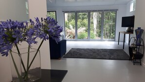 A modern living room with a large window, a blue sofa, a TV on a stand, and a vase with purple flowers. - Lucas Parklands - The Cube, peaceful getaway with direct access to 'Great Walk' (North Maleny)