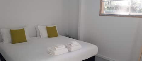Una cama bien hecha con sábanas blancas y dos toallas dobladas, un cabecero negro y una ventana con persianas.