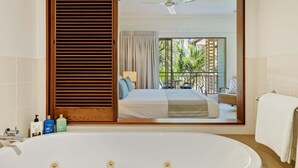 Een badkamer met een badkuip, een spiegel die een slaapkamer met een bed weerspiegelt, een plafondventilator en uitzicht op een balkon met planten.