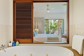Un bagno con una vasca, uno specchio che riflette una camera da letto con un letto, una ventola a soffitto e vista su un balcone con piante.
