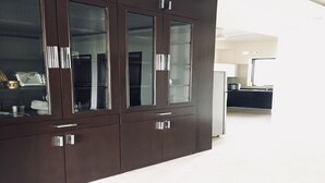 A dark wooden cabinet with glass doors and silver handles, located in a modern kitchen with white cabinets and a white refrigerator. - Moksha- Salvation (Gurugram)
