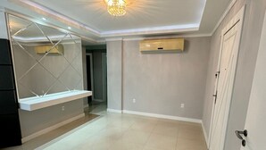Una habitación amplia con suelo de baldosas, una lámpara en el techo, aire acondicionado de pared y una puerta.