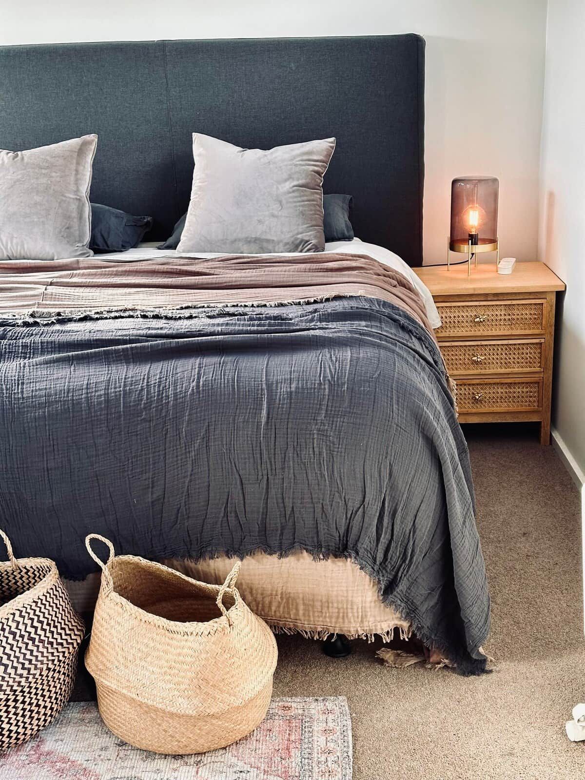 Un dormitorio con cabecera oscura, cama con ropa de cama gris, lámpara de noche y una cesta tejida en el suelo.