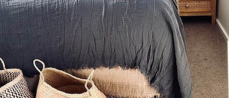 Un dormitorio con cabecera oscura, cama con ropa de cama gris, lámpara de noche y una cesta tejida en el suelo.