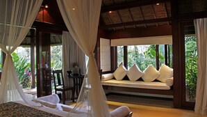 Ein Schlafzimmer mit Baldachinbett, einer Couch mit Kissen und Blick auf Grün hinter großen Fenstern.