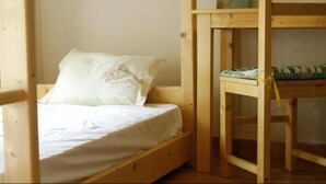 A single wooden bed with a white mattress and a floral pillow, a wooden table with a green cushion, and a wooden bunk bed structure. - Morabeza Kriol Hostel (Praia)