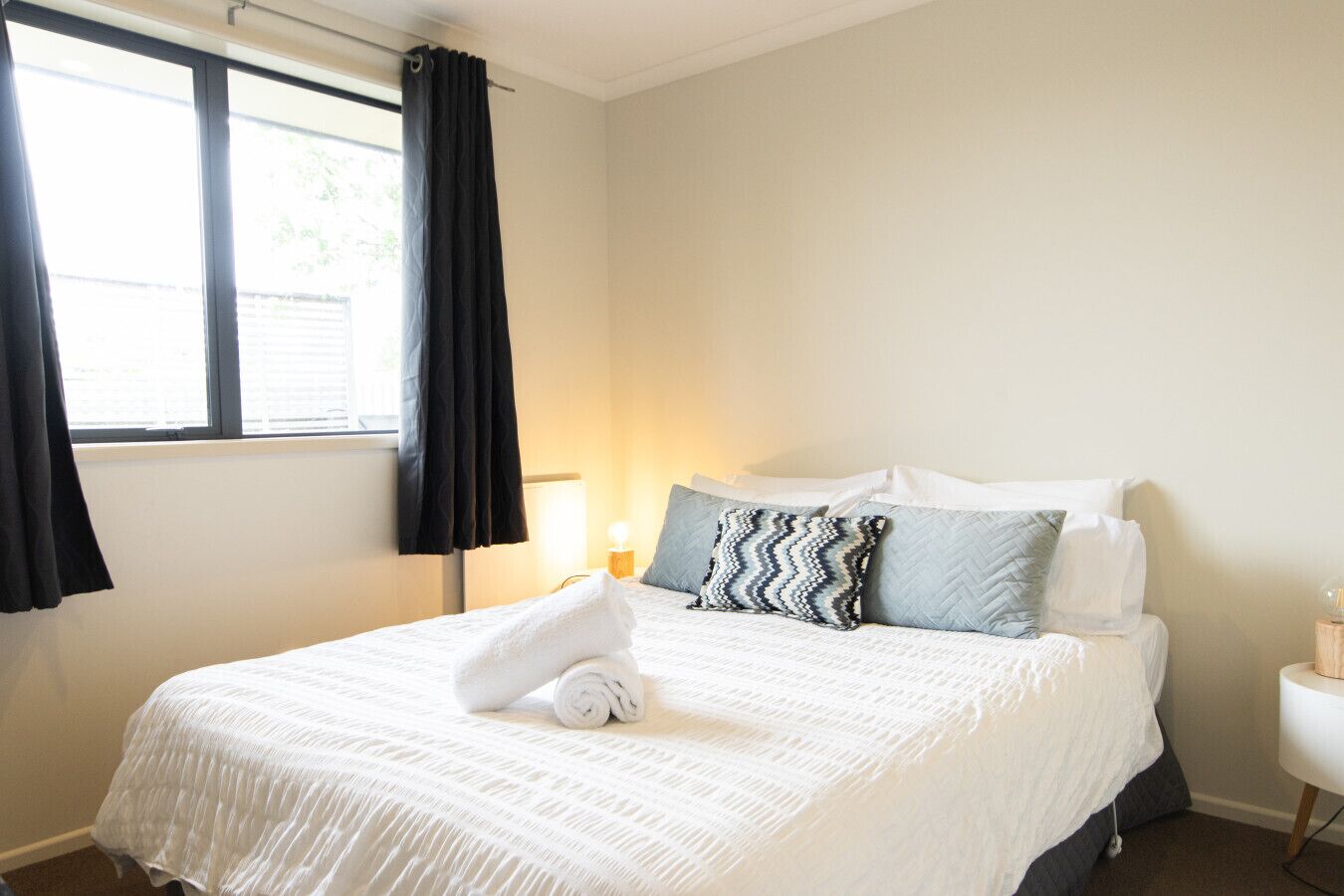 Habitación de hotel con cama, ventana con cortinas, lámpara de noche y una pequeña mesa con lámpara.