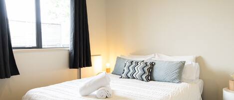 Habitación de hotel con cama, ventana con cortinas, lámpara de noche y una pequeña mesa con lámpara.