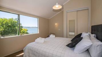 A bedroom with a large window, a bed with white bedding, a mirror, and a wall-mounted light fixture.