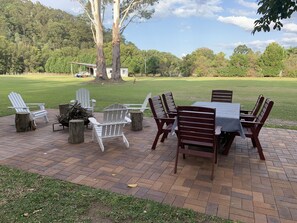 An outdoor patio with a brick floor, white Adirondack chairs, wooden chairs, and a table with a checkered tablecloth.