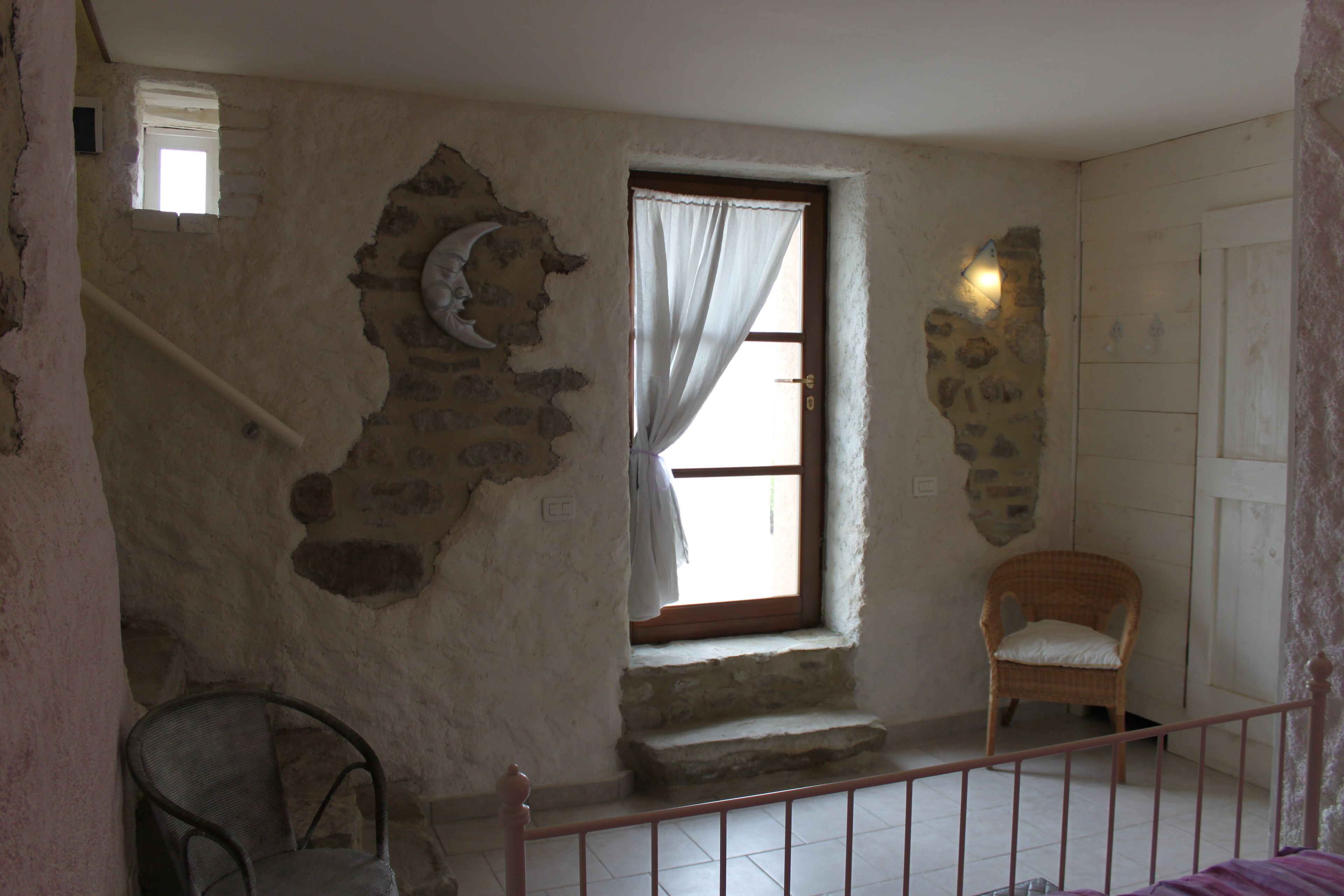 A room with a stone wall, a window with a curtain, a wicker chair, and a staircase with a railing.