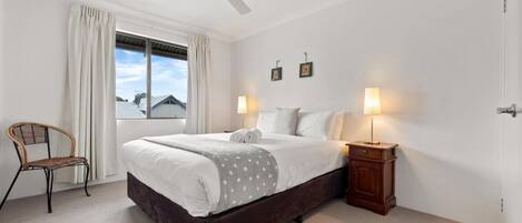 A bedroom with a bed, a chair, a nightstand, a ceiling fan, and a window with curtains.